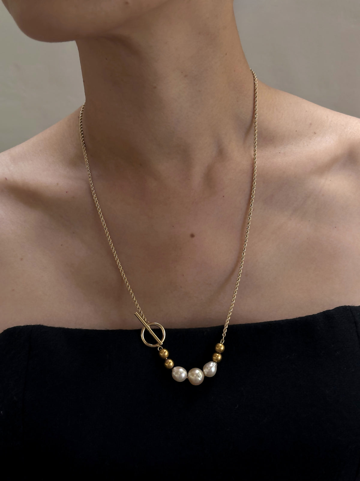 A woman wears the Trio of Vintage Pearls and 14K Beads on a 14K Gold Chain with Toggle Closure, featuring alternating vintage pearls and gold beads. The T-bar toggle necklace graces her collarbone above a black strapless top against a neutral backdrop.