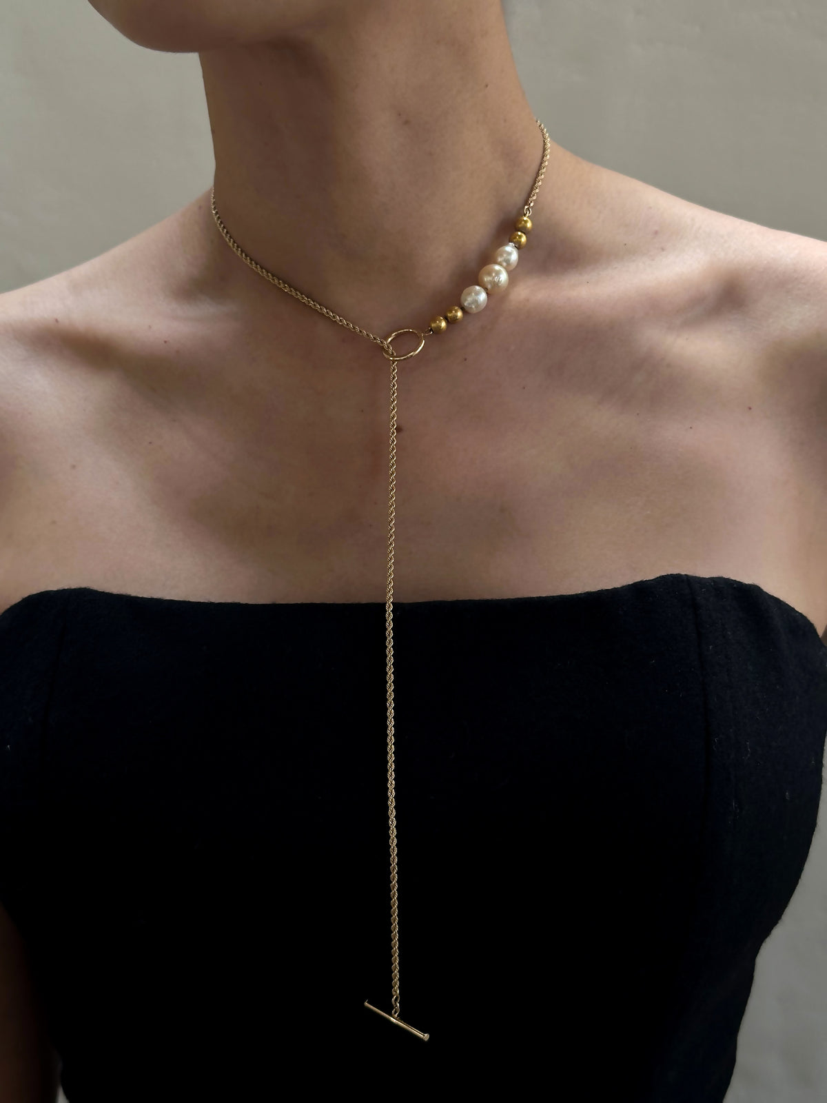 A person in a strapless black top displays the "Trio of Vintage Pearls and 14K Beads on 14K Gold Chain with Toggle Closure," a striking gold lariat necklace. The asymmetrical design features vintage pearls and gold beads on one side, with an elegant, long chain gracefully hanging down.