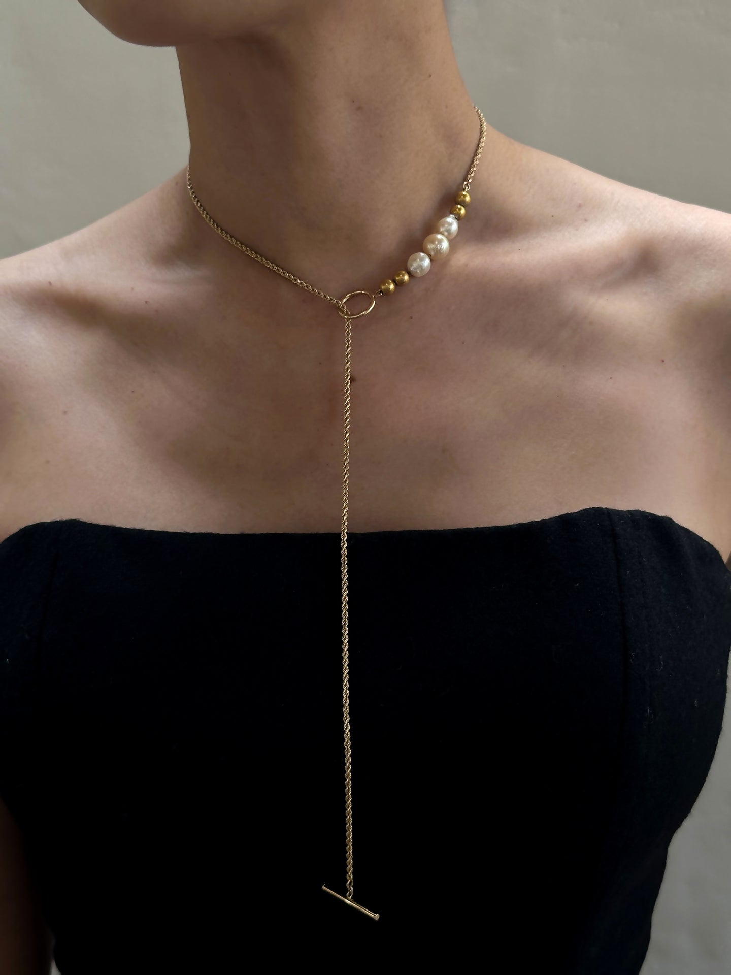 A person wearing a strapless black top and a Vintage 14K Gold Chain with Trio of Vintage Baroque Pearls and 14K Beads c.1950, featuring a long, thin 14K gold twisted rope chain that hangs down the front, ending in a bar pendant. The background is plain and neutral.