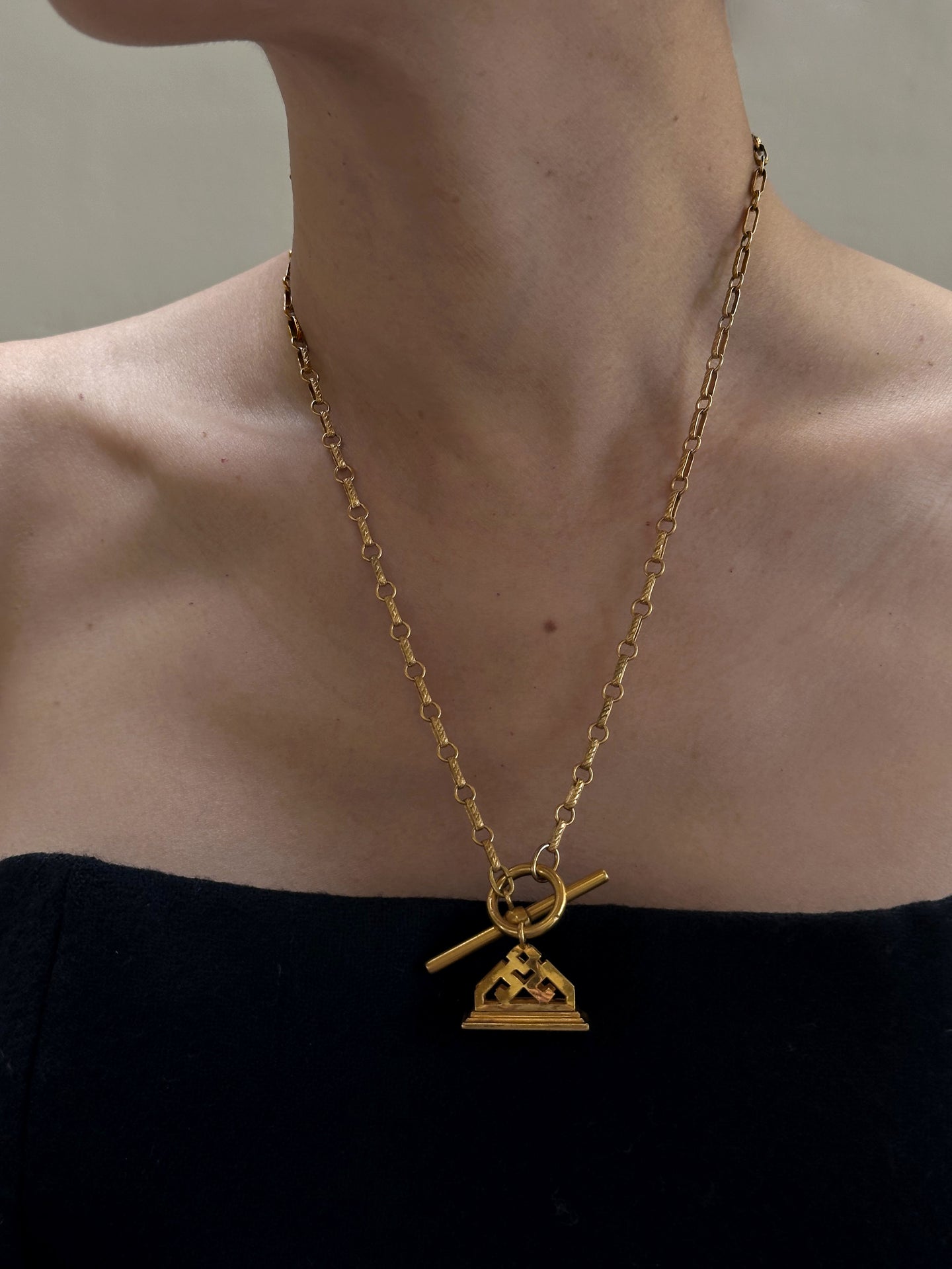 A person wearing a black strapless top showcases the Antique Gold Filled Fob and Chain with Front Closing T-bar c.1910-1930. This exquisite necklace features a toggle clasp and a pendant with a geometric design, reminiscent of classic fob and chain styles. The neutral background keeps the focus on this elegant jewelry piece.