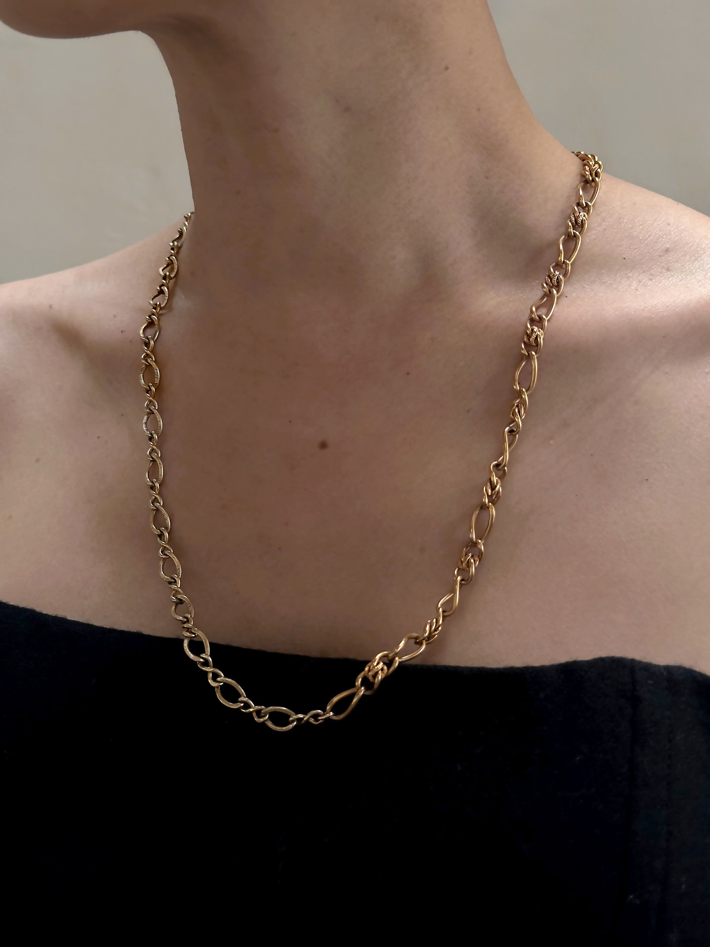 A person wearing an Antique Gold Filled Twisted Curb Chain necklace, dating circa 1890 and 1940, with a series of interlocking links. The person is dressed in an off-the-shoulder black garment, revealing their neck, collarbone, and upper chest. The background is neutral and out of focus.