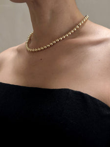 A close-up of a person wearing the Lan Jaenicke Signature 14k Gold Bead Necklace. The person is dressed in an off-the-shoulder black garment, showcasing the elegant simplicity of the jewelry against the bare skin of their neck and shoulders. The background is neutral and out of focus.