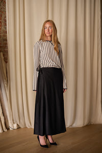 A woman with long hair is standing in front of a beige curtain, wearing a handmade white Qipao Blouse | Crepe de Chine Custom Houndstooth with a geometric pattern and a long black skirt, paired with black high heels. The background features a brick wall on the left.