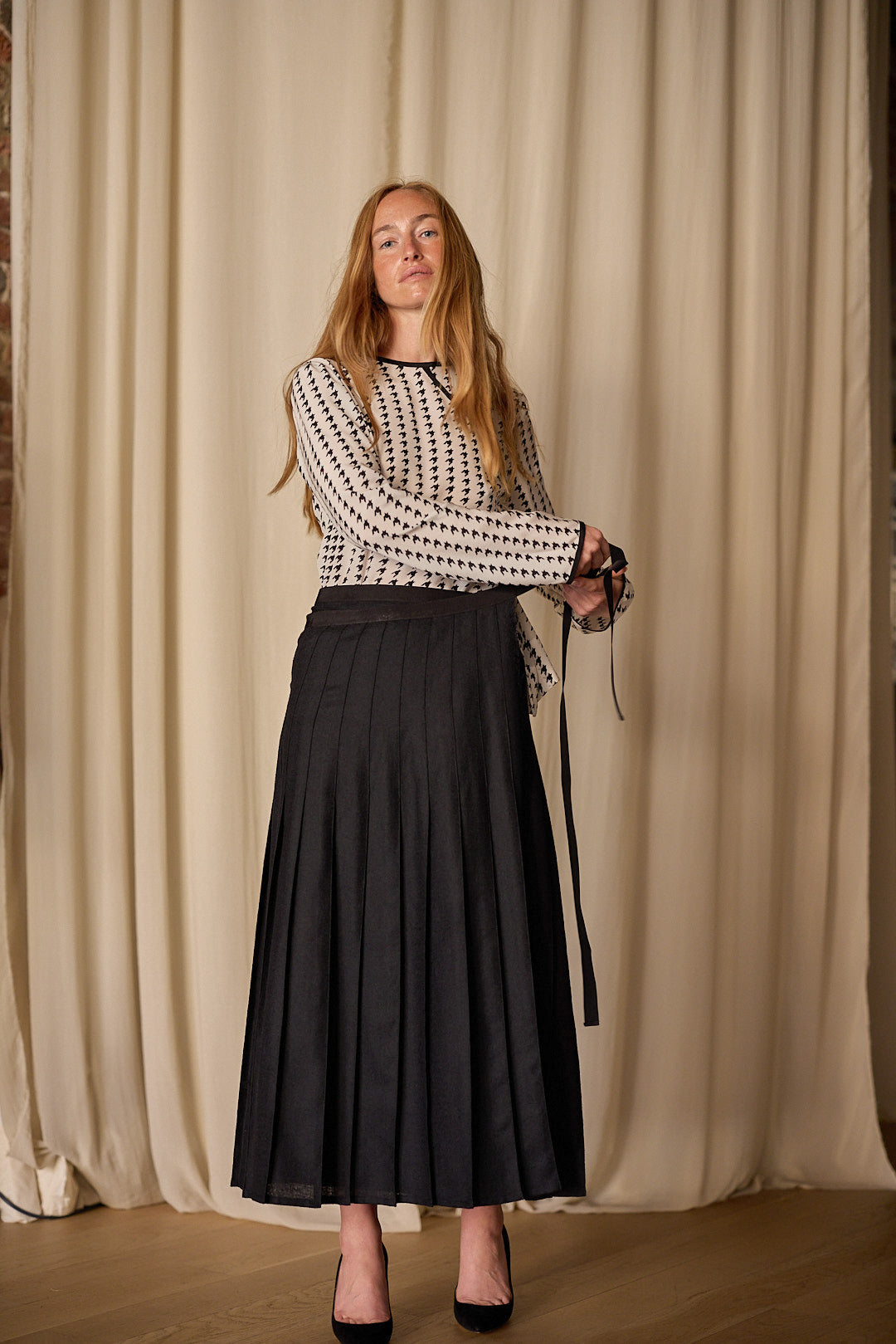 A person with long red hair stands in front of beige curtains, wearing a patterned long-sleeve shirt and the Hand Pleated Wrap Skirt made from Japanese wool gauze. This zero-waste design adds elegance as they hold a black ribbon and wear matching black shoes.