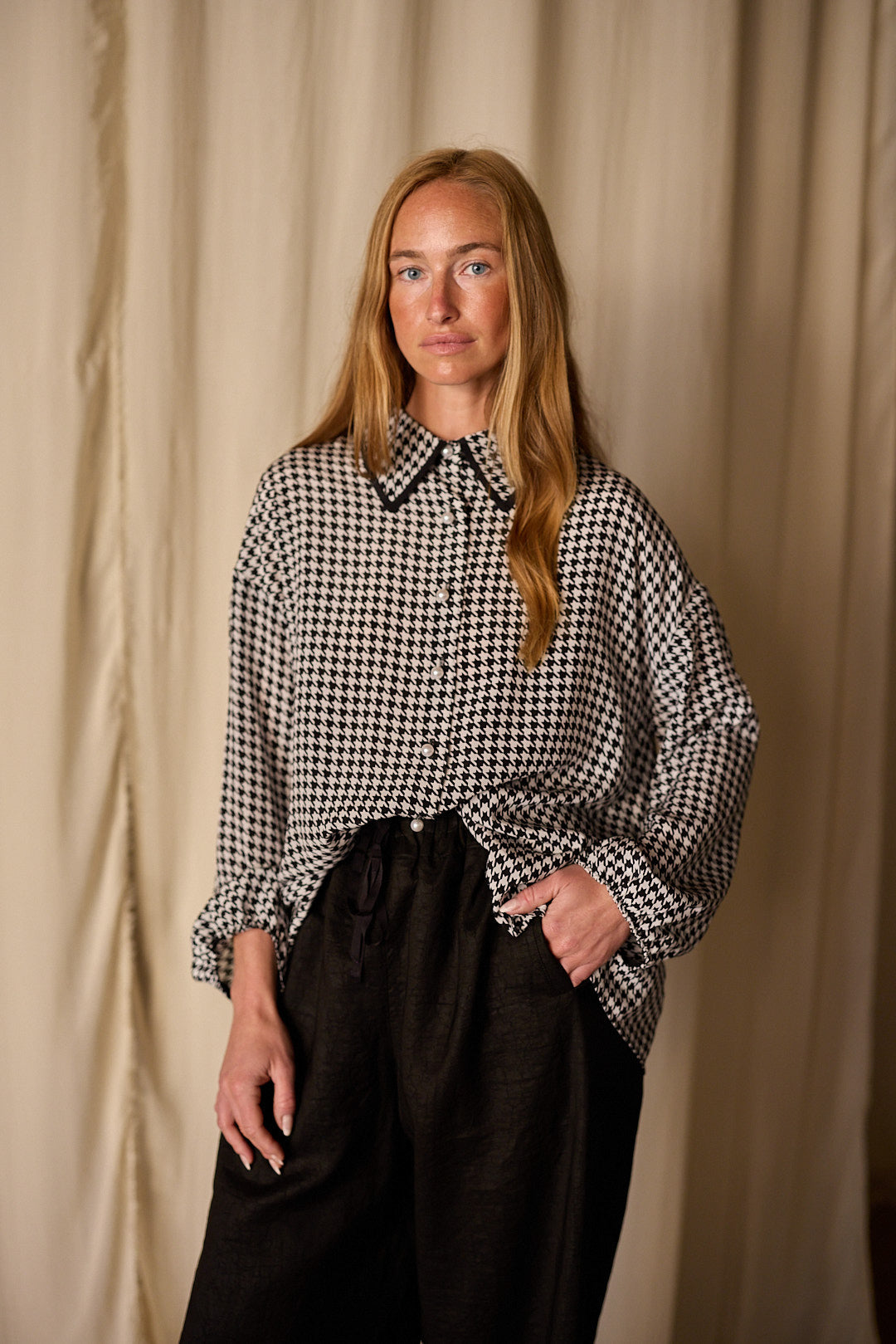 A person with long, straight hair stands in front of a neutral-colored curtain, wearing a black-and-white houndstooth patterned Painter's Shirt | Crepe de Chine Houndstooth and dark trousers by Atelier Lan Jaenicke. Made from 100% silk crepe de chine, they have one hand in their pocket and a relaxed expression, gazing directly at the camera.