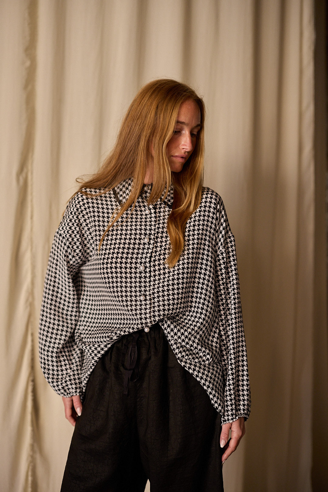 A woman with long, light brown hair is standing against a beige curtain backdrop. She is wearing a black and white houndstooth patterned Painter's Shirt | Crepe de Chine Houndstooth made of 100% silk crepe de chine and black pants. Her gaze is directed downwards and to the left.