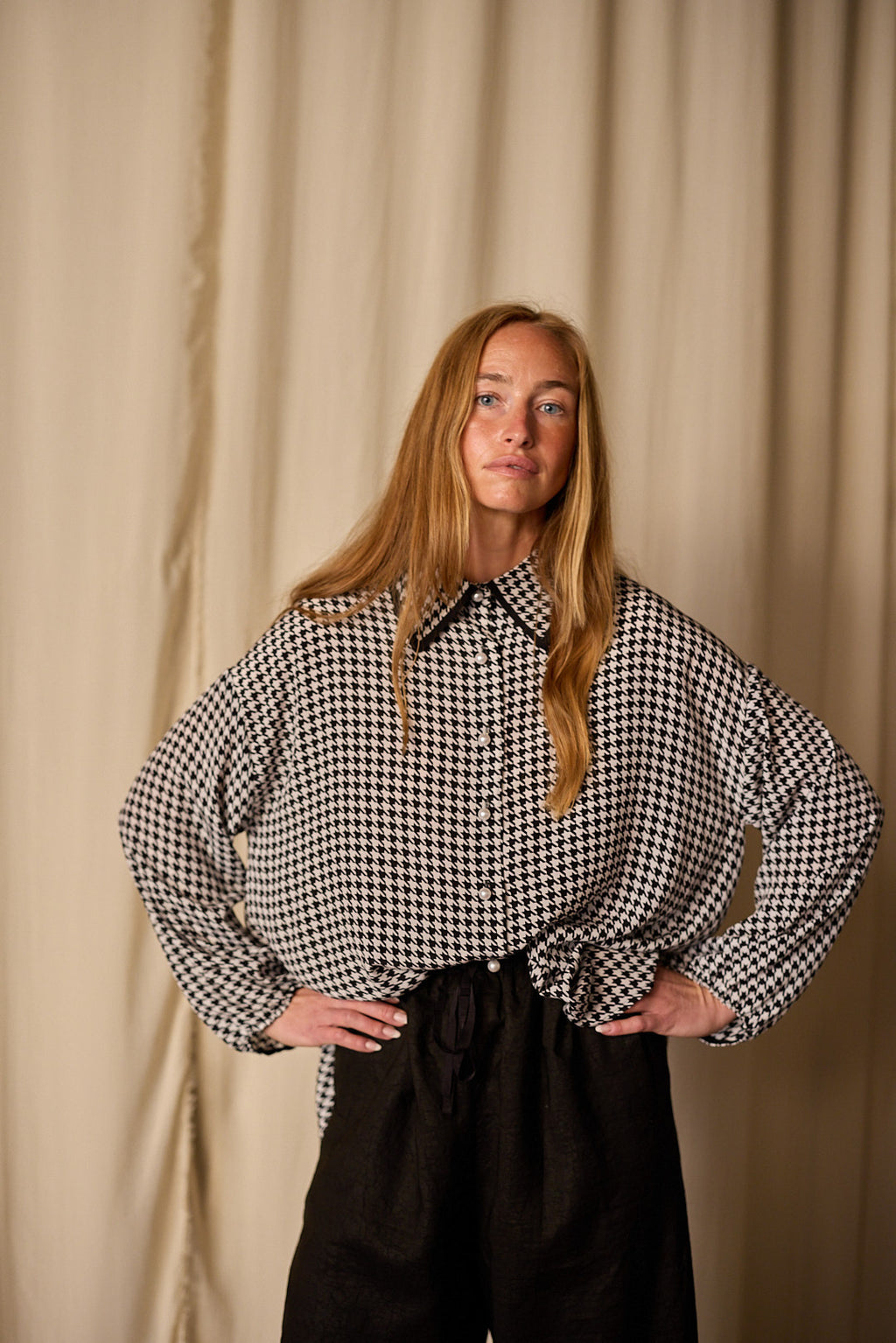 A woman with long blonde hair stands in front of a beige curtain. She is wearing Atelier Lan Jaenicke's Painter's Shirt | Crepe de Chine Houndstooth, crafted from 100% silk crepe de chine, tucked into black pants. Her hands are resting on her hips.