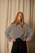 A woman with long blonde hair stands in front of a beige curtain. She is wearing Atelier Lan Jaenicke's Painter's Shirt | Crepe de Chine Houndstooth, crafted from 100% silk crepe de chine, tucked into black pants. Her hands are resting on her hips.