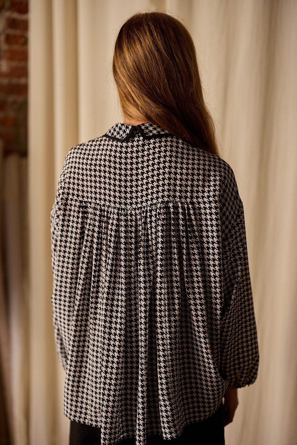A person with long, light brown hair is standing with their back to the camera, wearing a black and white houndstooth patterned Painter's Shirt | Crepe de Chine Houndstooth with a pleated back. The blouse, 100% silk crepe de chine from Atelier Lan Jaenicke, contrasts beautifully against the light-colored curtain and partial brick wall.