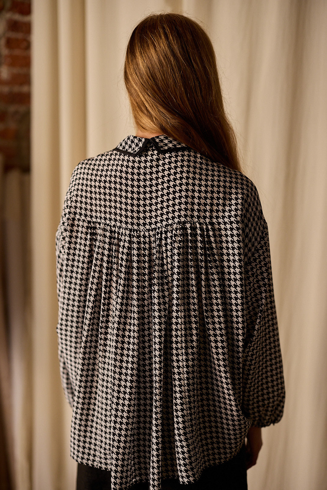 A person with long, light brown hair is standing with their back to the camera, wearing a black and white houndstooth patterned Painter's Shirt | Crepe de Chine Houndstooth with a pleated back. The blouse, 100% silk crepe de chine from Atelier Lan Jaenicke, contrasts beautifully against the light-colored curtain and partial brick wall.