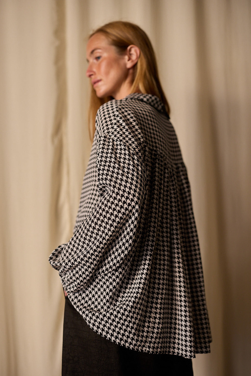 A person with long blond hair is captured in profile, wearing a loose-fitting, Painter's Shirt | Crepe de Chine Houndstooth. They are facing to the left in front of a plain, light-colored curtain background. The elegant top is crafted from 100% silk crepe de chine by Atelier Lan Jaenicke.