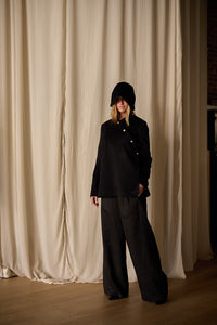 A person stands in front of beige curtains, wearing a loose-fitting Qipao Jacket | Cashmere Black with gold buttons and dark wide-legged pants. This elegant ensemble by Atelier Lan Jaenicke features a handmade touch. Their hands are in their pockets, and their face is partially obscured by the hood's shadow.