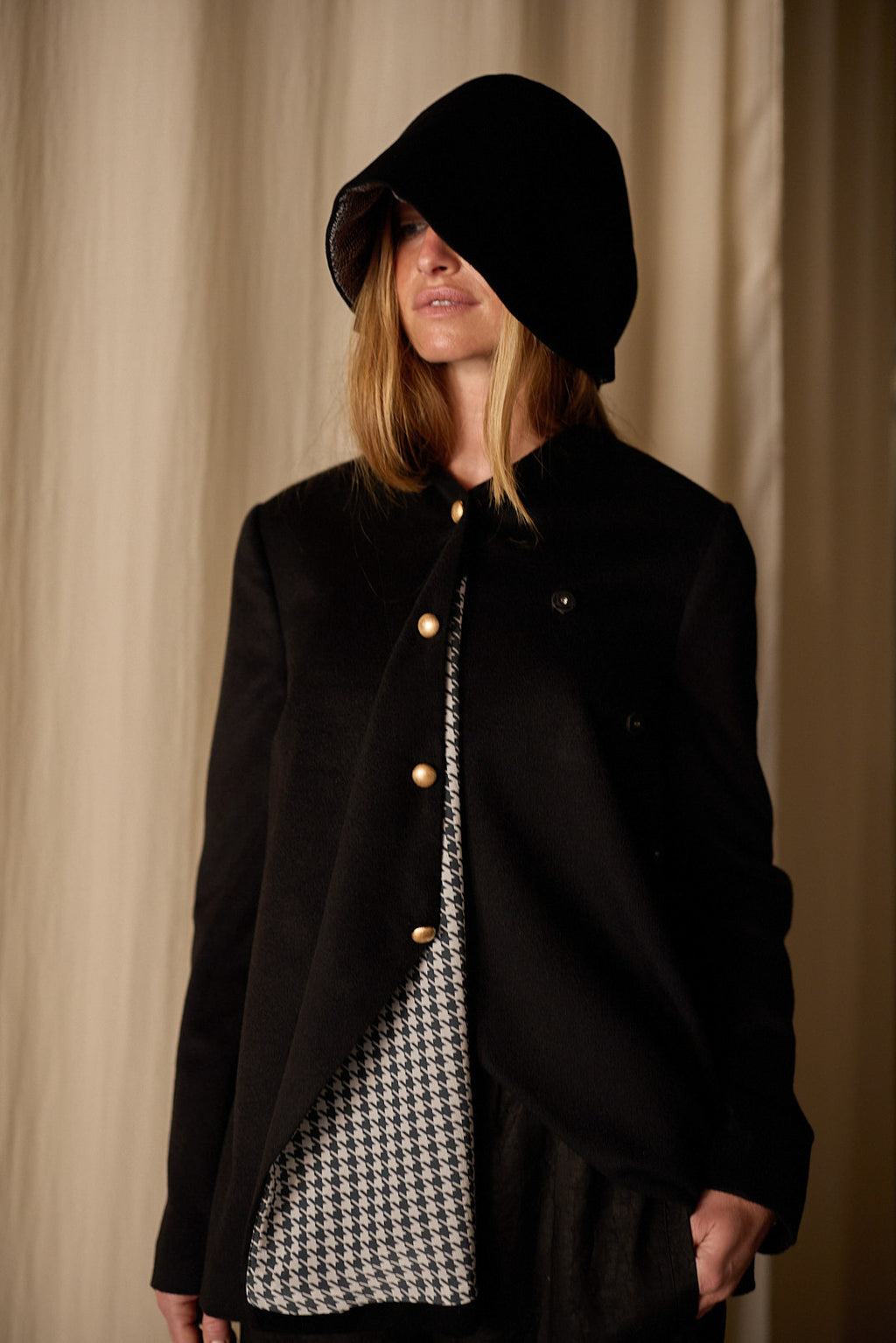 A person wearing a large black hooded coat partially covering their face. The Qipao Jacket | Cashmere Black, an exquisite Atelier Lan Jaenicke creation, is open to reveal a houndstooth-patterned inner layer. The background consists of beige curtains.