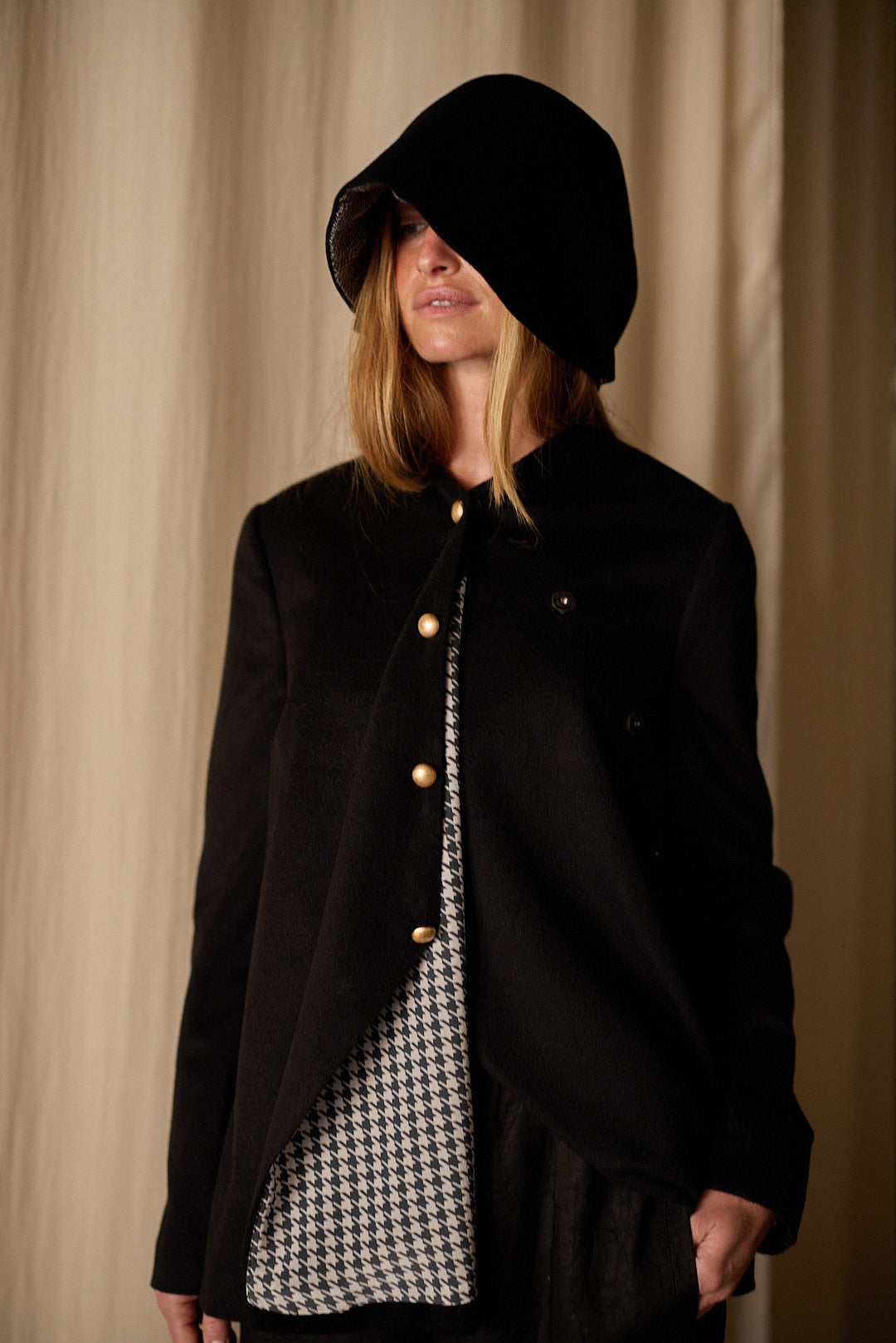 A person wearing a large black hooded coat partially covering their face. The Qipao Jacket | Cashmere Black, an exquisite Atelier Lan Jaenicke creation, is open to reveal a houndstooth-patterned inner layer. The background consists of beige curtains.