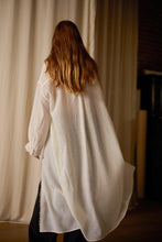 Load image into Gallery viewer, A person with long, reddish-brown hair stands with their back to the camera, wearing a loose, white Dress Coat | Japanese Wool Gauze Undyed. The background is softly lit with neutral-colored curtains and some dark furniture partially visible, evoking a sense of sustainable design.