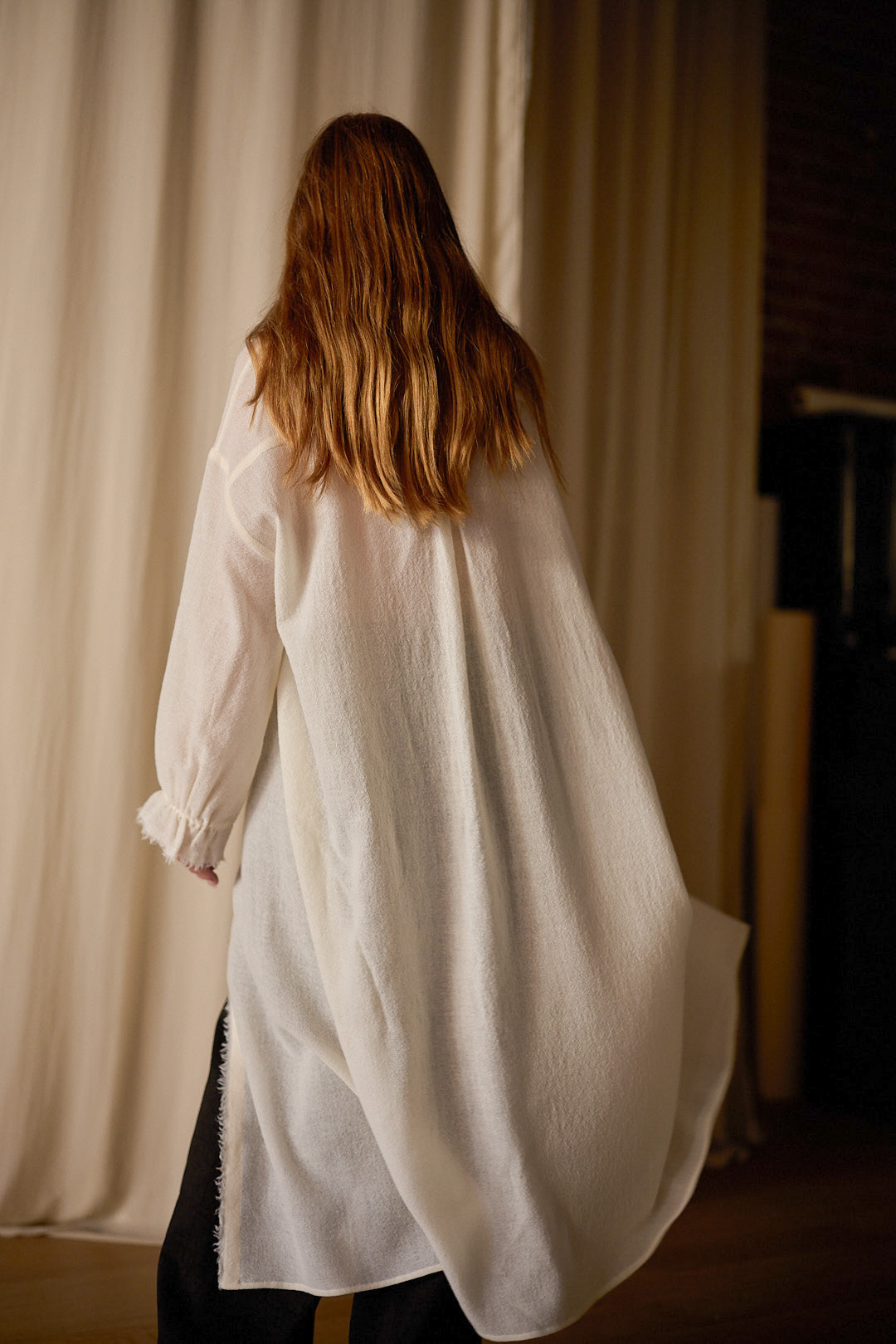A person with long, reddish-brown hair stands with their back to the camera, wearing a loose, white Dress Coat | Japanese Wool Gauze Undyed. The background is softly lit with neutral-colored curtains and some dark furniture partially visible, evoking a sense of sustainable design.