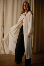 Load image into Gallery viewer, A person stands indoors wearing a long, flowing Dress Coat | Japanese Wool Gauze Undyed over a pair of loose black pants. The jacket, featuring sustainable design with a tie at the collar, drapes elegantly to the floor. Cream-colored curtains provide a soft background.