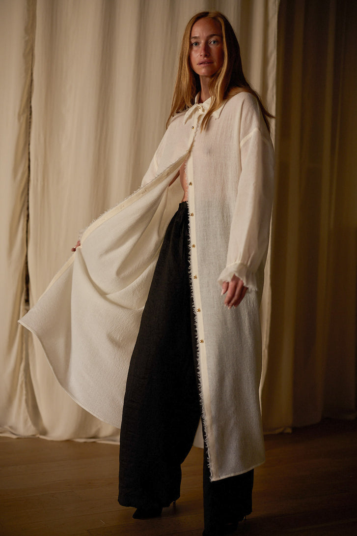 A person stands indoors wearing a long, flowing Dress Coat | Japanese Wool Gauze Undyed over a pair of loose black pants. The jacket, featuring sustainable design with a tie at the collar, drapes elegantly to the floor. Cream-colored curtains provide a soft background.