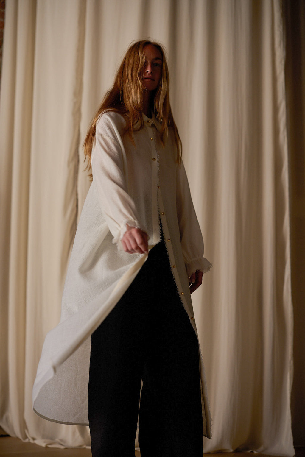 A person with long hair stands against a backdrop of flowing beige curtains. They are wearing a Dress Coat | Japanese Wool Gauze Undyed over black pants. The coat flares out slightly at the bottom as they hold it with one hand, while looking towards the camera.