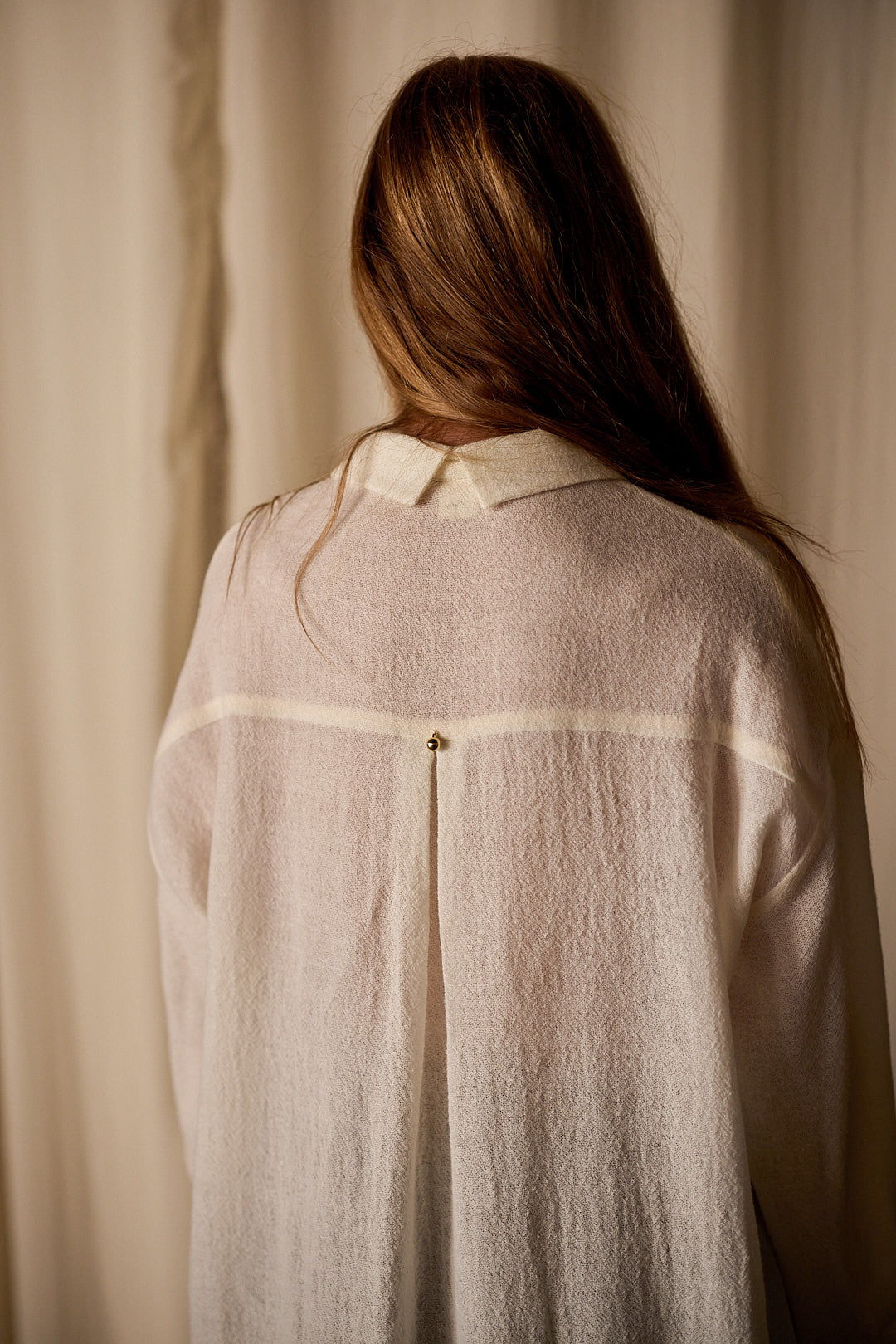 A person with long brown hair is seen from behind, wearing a sheer, white Dress Coat | Japanese Wool Gauze Undyed. The focus is on the back of the dress coat, which features a small gold-colored pin or button at the center. The background is neutral with light-colored curtains, highlighting sustainable design.