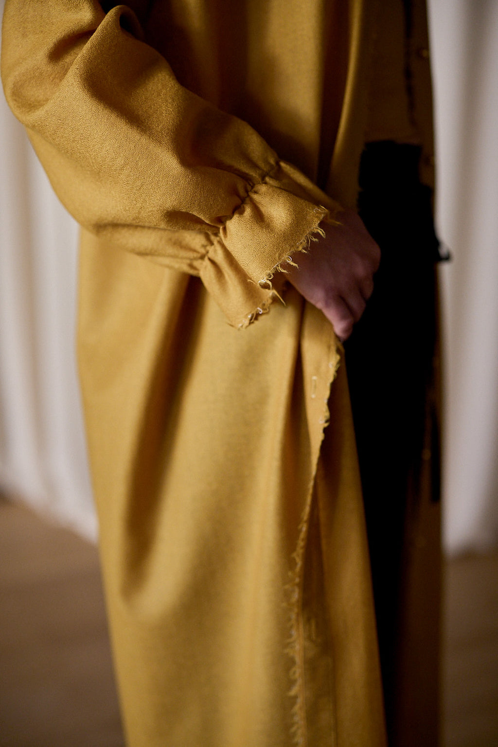 Close-up of a person wearing the Dress Coat | Japanese Wool Gauze Gold with frayed edges, crafted from Japanese wool gauze. The person is holding the coat's lapel with one hand. The sleeve has gathered fabric at the wrist, adding a textured detail. The background is softly blurred, emphasizing the handmade in-house garment.