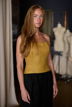 Load image into Gallery viewer, A woman with long reddish-brown hair stands in a room adorned with dressmaker&#39;s mannequins. She wears a Custom Bustier | Japanese Wool Gauze Gold and a black skirt. The room features dark and light curtains, mirrors, and Japanese wool gauze fabrics, all handmade in-house, creating an elegant atmosphere.