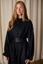 Load image into Gallery viewer, A woman with long, straight blonde hair is standing in front of a neutral-colored backdrop. She is wearing a black Dress Coat | Japanese Wool Gauze Black with a button-up front and a black belt at the waist. The dress coat, handmade in-house, complements her calm demeanor as she looks directly at the camera.