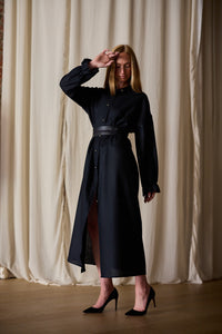 A woman stands indoors in front of beige curtains, wearing a Dress Coat | Japanese Wool Gauze Black with a belt at the waist. She has one hand near her face and the other resting by her side. She is wearing black high heels and has straight, shoulder-length hair.