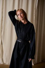 Load image into Gallery viewer, A woman with long blonde hair stands in front of beige curtains. She is wearing a Dress Coat | Japanese Wool Gauze Black with a belt at the waist. Her left hand is placed on top of her head as she gazes towards the camera with a neutral expression.