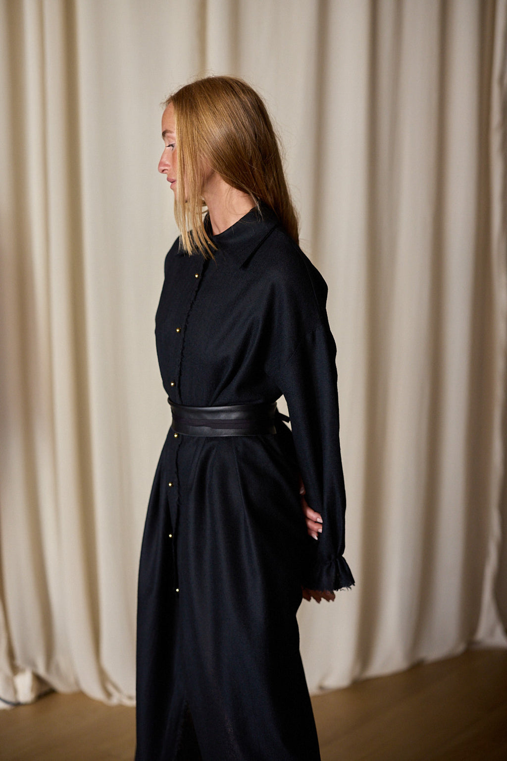 A woman with long blonde hair is standing in profile against a backdrop of beige curtains. She is dressed in a Dress Coat | Japanese Wool Gauze Black, with buttons down the front and a wide black belt. Her arms are positioned behind her back, exuding an air of timeless elegance.
