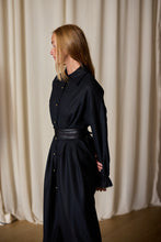 Load image into Gallery viewer, A woman with long blonde hair is standing in profile against a backdrop of beige curtains. She is dressed in a Dress Coat | Japanese Wool Gauze Black, with buttons down the front and a wide black belt. Her arms are positioned behind her back, exuding an air of timeless elegance.