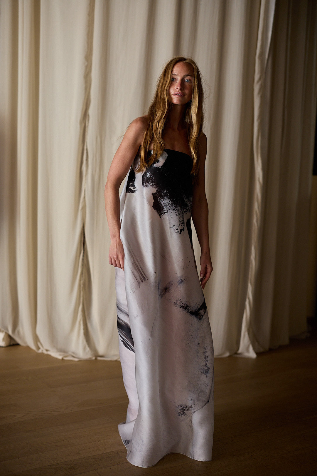 A woman with long hair stands in a softly lit room, wearing a sleeveless, floor-length Custom Shuimo Painting Dress | Satin Organza Buckhouse Print. Beige curtains and wooden flooring are visible in the background, creating a minimalistic and serene atmosphere.
