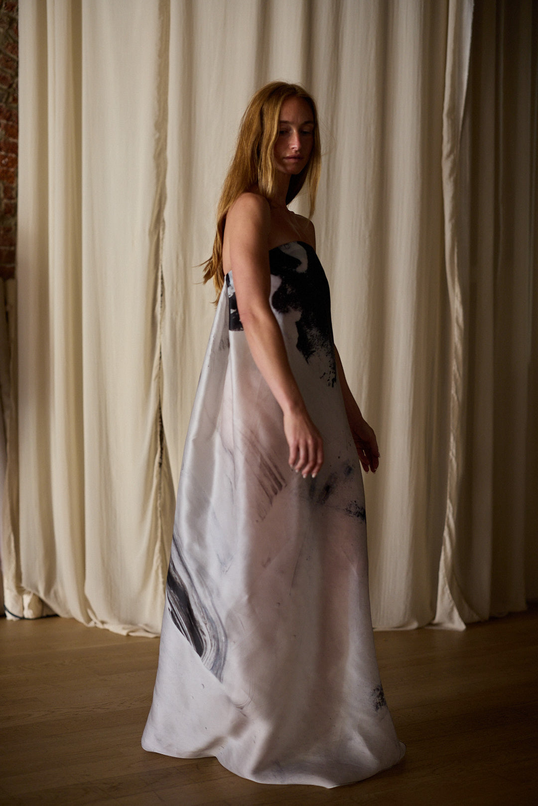 A woman with long, wavy hair poses in a Custom Shuimo Painting Dress | Satin Organza Buckhouse Print featuring an exclusive print with a monochromatic abstract pattern. She stands in front of light-colored curtains, with soft, warm lighting creating a gentle ambiance.