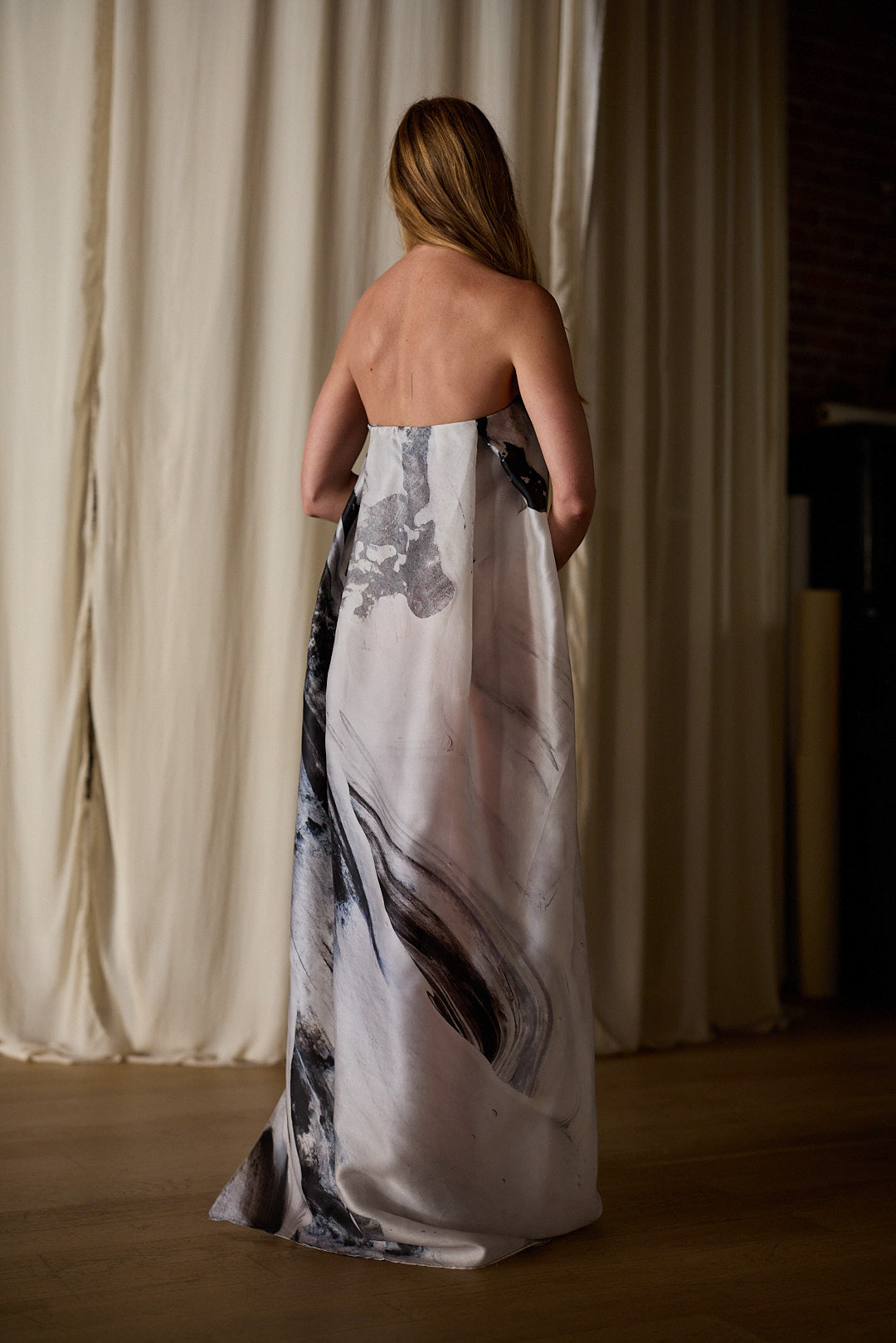 A woman with long hair stands with her back to the camera, wearing a Custom Shuimo Painting Dress | Satin Organza Buckhouse Print. She is in a dimly lit room with cream-colored curtains as the background.