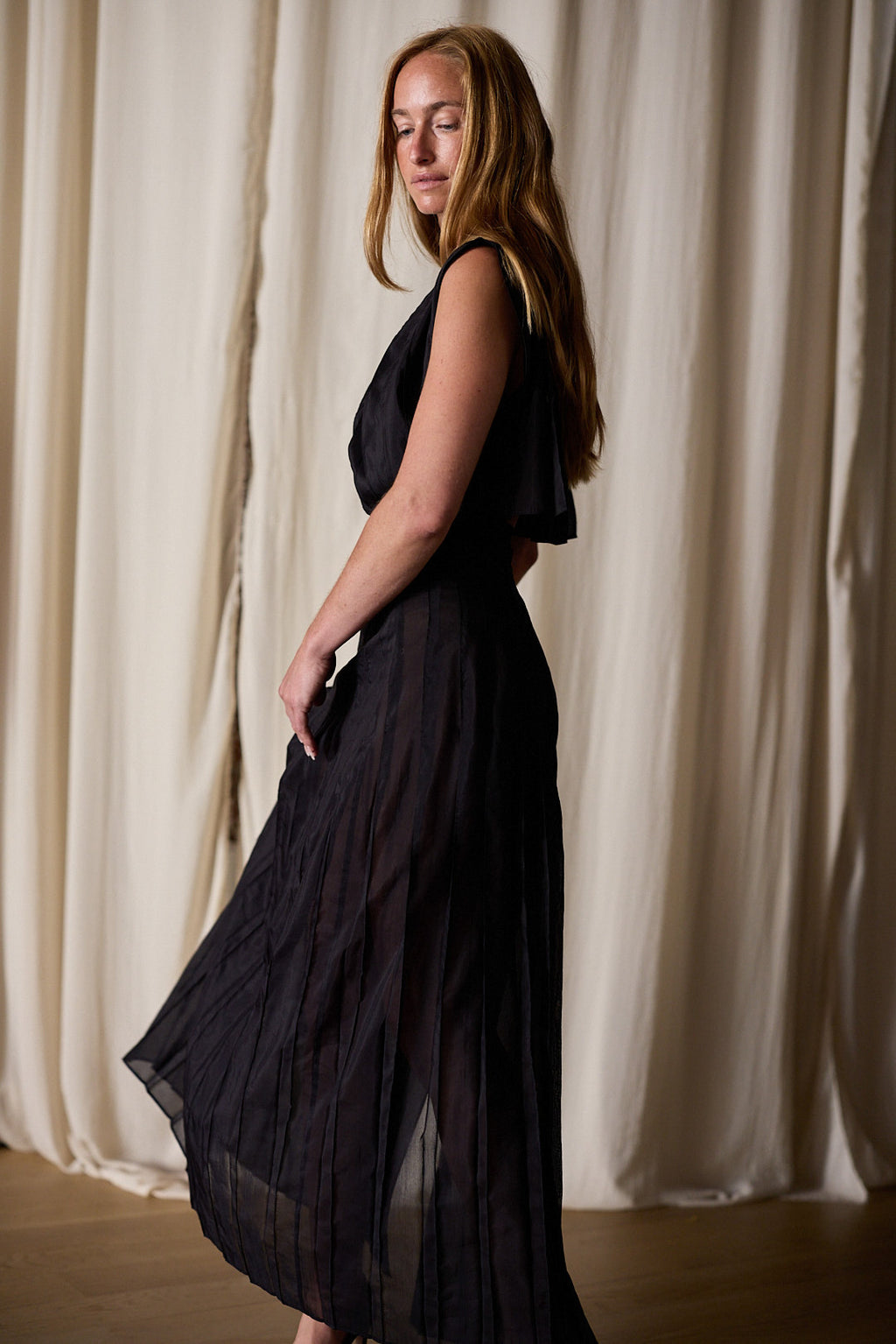 A woman with long, red hair stands in profile, wearing a Hand Pleated Column Dress | Black Cotton Silk Voile. She poses gracefully in front of beige, floor-length curtains, with her eyes closed, giving a serene and reflective expression.