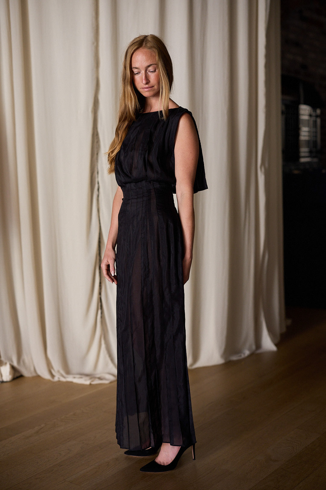 A woman with long, straight hair is wearing the Hand Pleated Column Dress | Black Cotton Silk Voile. She stands indoors on a wooden floor in front of cream-colored draped curtains, looking down with her arms relaxed at her sides. She has one leg slightly bent, wearing black heels.