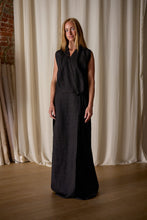 Load image into Gallery viewer, A person stands in a well-lit room with beige curtains and a brick wall in the background. They wear a long, sleeveless, dark Xiang Yun Silk Wrap Dress with wrap detail and buttons down the front. Crafted from Xiang Yun Silk and handmade in-house, it embodies sustainable design. The person has light brown hair and a relaxed, confident posture.