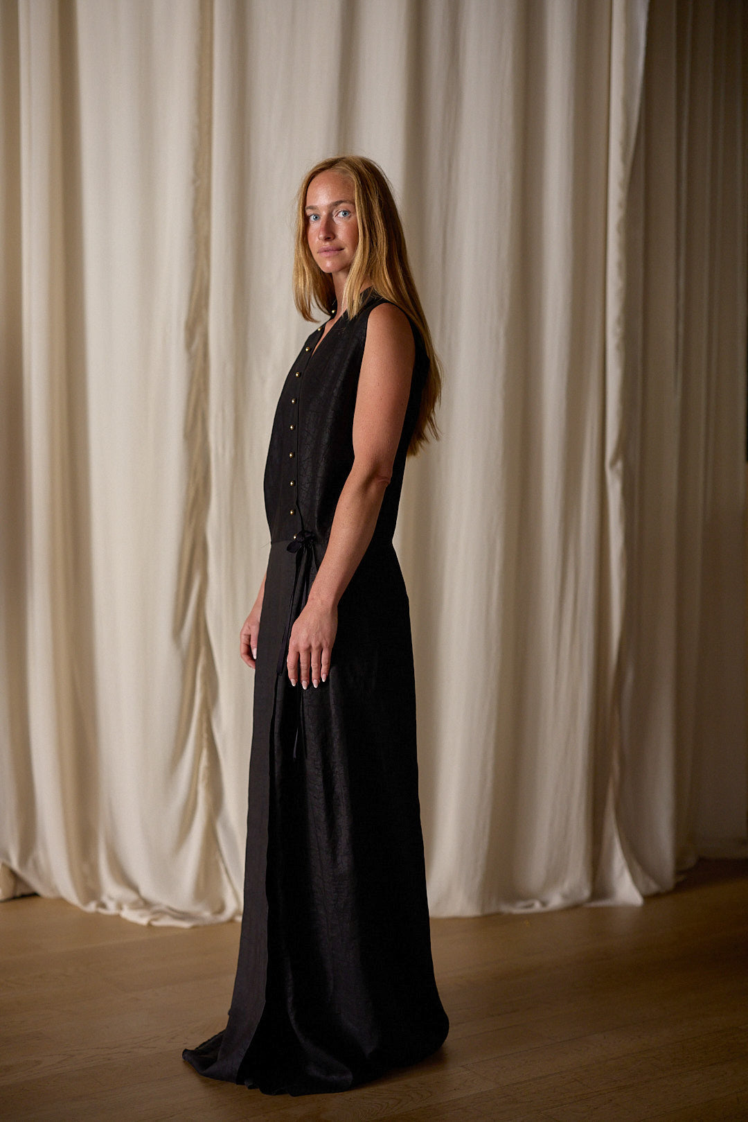 A person with long hair stands sideways, looking at the camera, in front of a light-colored curtain. They are wearing a sleeveless, long black Xiang Yun Silk Wrap Dress and have a neutral expression on their face. The floor is wooden.