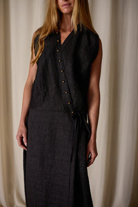 A person with long blonde hair is wearing a Xiang Yun Silk Wrap Dress, handmade in-house, featuring a textured fabric and a row of buttons on one side. The background consists of white fabric drapes. The person's face is not fully visible.