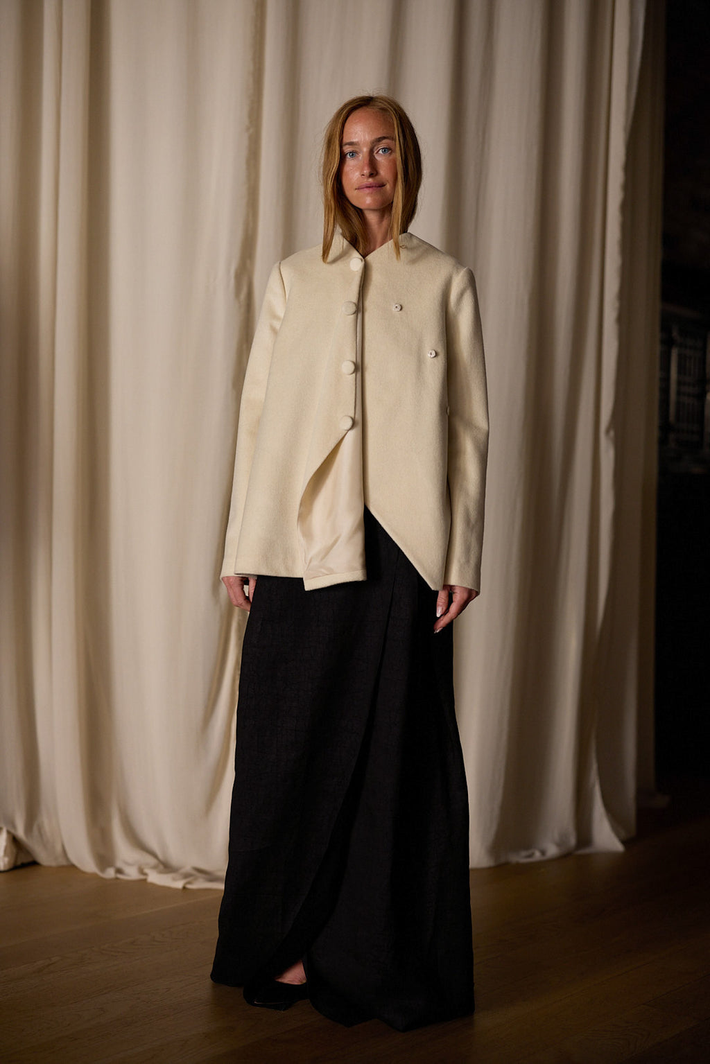 A person with straight, mid-length hair stands indoors in front of beige curtains. They are wearing a stylish cream-colored, handmade Qipao Jacket | Cashmere Undyed with an asymmetrical design and large buttons, paired with a long, dark skirt. The jacket features an undyed silk lining. They have a neutral expression and hands by their sides.