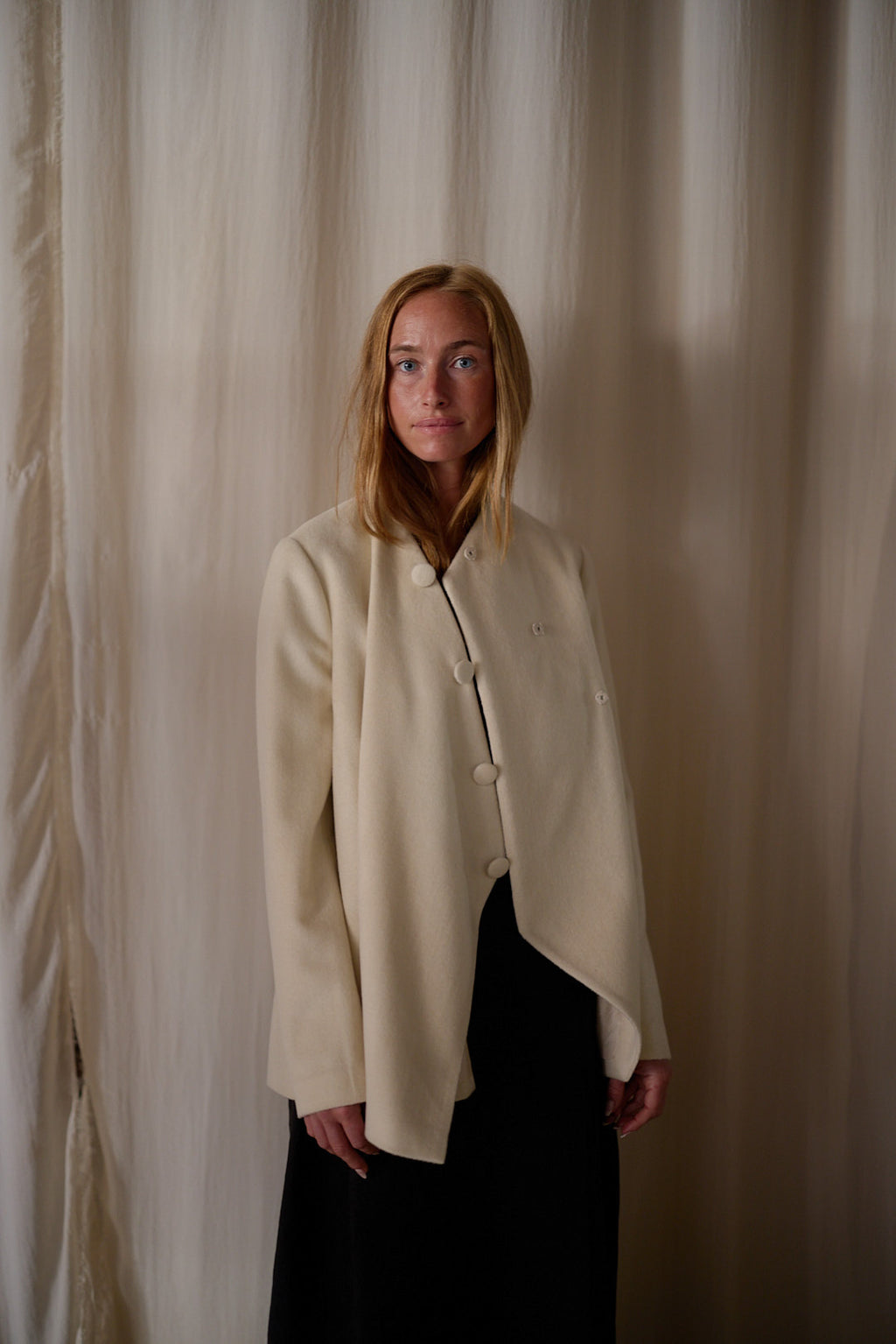 A woman with long blonde hair stands in front of beige curtains. She is wearing a sophisticated cream-colored Qipao Jacket | Cashmere Undyed, featuring an undyed silk lining and buttoned at the top while draping open at the bottom, paired with a dark skirt. The Qipao Jacket | Cashmere Undyed is handmade in-house, and she has a composed expression on her face.