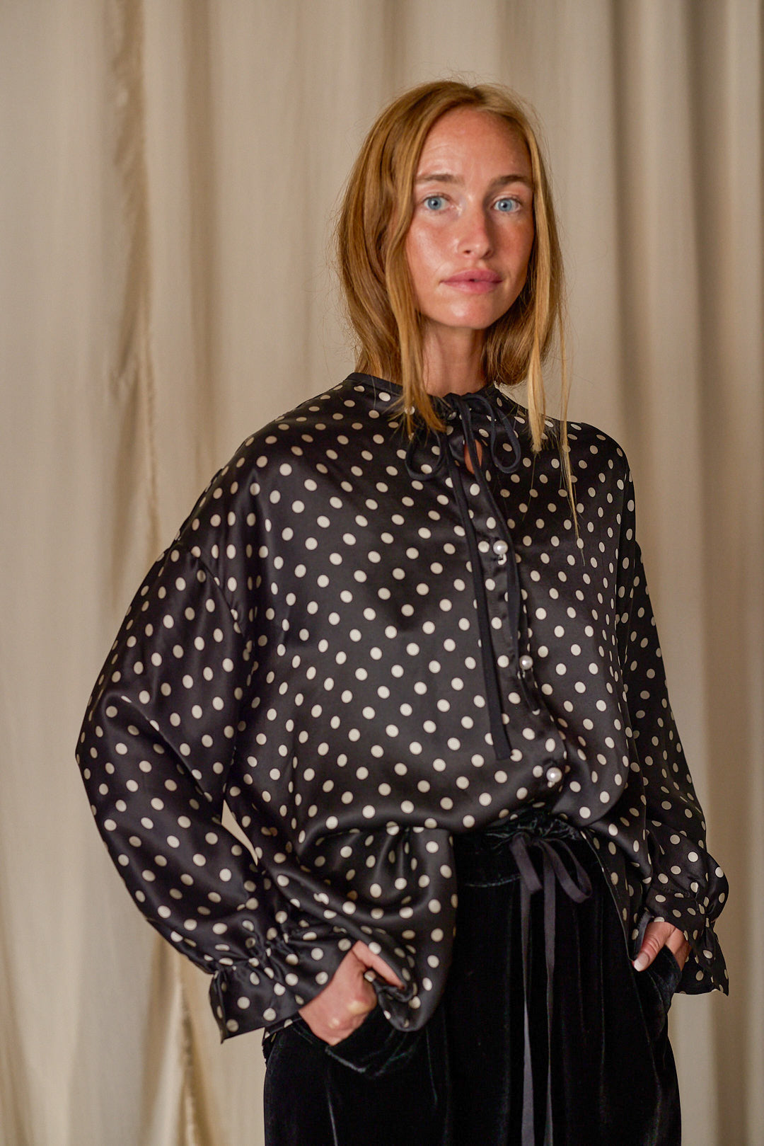 A person with long blonde hair is standing in front of a beige curtain, wearing a Ribbon Shirt | Black Polkadot Silk Charmeuse. The blouse has a bow tied at the neckline and voluminous sleeves, evoking the elegance of silk charmeuse. The person exudes a calm and thoughtful expression.