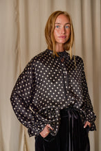Load image into Gallery viewer, A person with long blonde hair is standing in front of a beige curtain, wearing a Ribbon Shirt | Black Polkadot Silk Charmeuse. The blouse has a bow tied at the neckline and voluminous sleeves, evoking the elegance of silk charmeuse. The person exudes a calm and thoughtful expression.