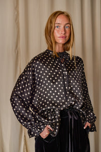A person with long blonde hair is standing in front of a beige curtain, wearing a Ribbon Shirt | Black Polkadot Silk Charmeuse. The blouse has a bow tied at the neckline and voluminous sleeves, evoking the elegance of silk charmeuse. The person exudes a calm and thoughtful expression.
