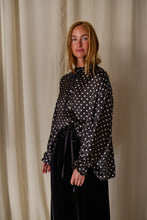 Load image into Gallery viewer, A person with long blonde hair stands in front of a light-colored curtain, smiling. They are wearing a Ribbon Shirt | Black Polkadot Silk Charmeuse and long sleeves tied at the wrists, paired with dark pants.