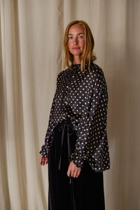 A person with long blonde hair stands in front of a light-colored curtain, smiling. They are wearing a Ribbon Shirt | Black Polkadot Silk Charmeuse and long sleeves tied at the wrists, paired with dark pants.