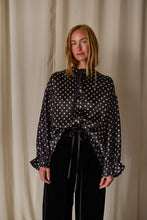 Load image into Gallery viewer, A woman with long blonde hair stands against a beige curtain backdrop. She is wearing a handmade, long-sleeved black blouse with white polka dots, paired with black pants. The Ribbon Shirt | Black Polkadot Silk Charmeuse features a tied neck and ruffled cuffs at the sleeves.