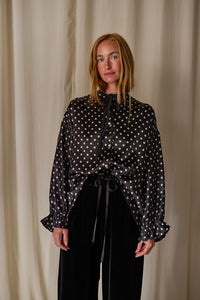 A woman with long blonde hair stands against a beige curtain backdrop. She is wearing a handmade, long-sleeved black blouse with white polka dots, paired with black pants. The Ribbon Shirt | Black Polkadot Silk Charmeuse features a tied neck and ruffled cuffs at the sleeves.