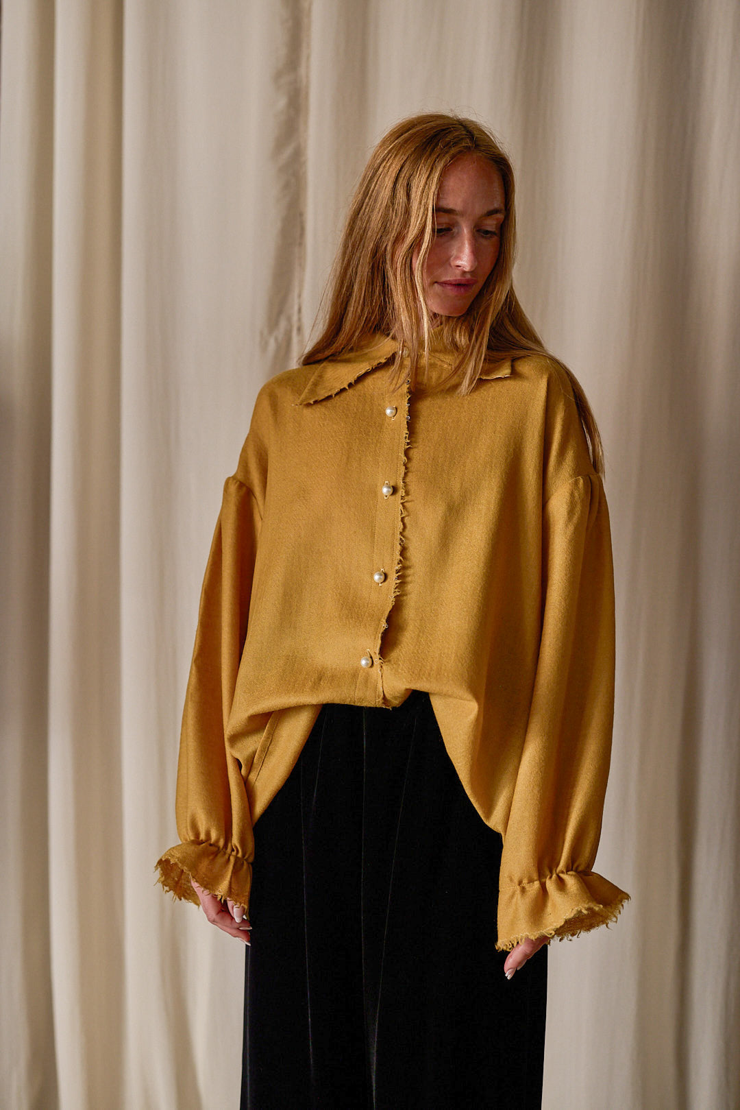 A person with long, light brown hair is standing against a light-colored curtain background. They are wearing a mustard yellow Poet Shirt | Japanese Wool Gauze Gold with pearl buttons and frilled cuffs, paired with black pants. The blouse, made from Japanese wool gauze, has a distinct collar and a loose, oversized fit.