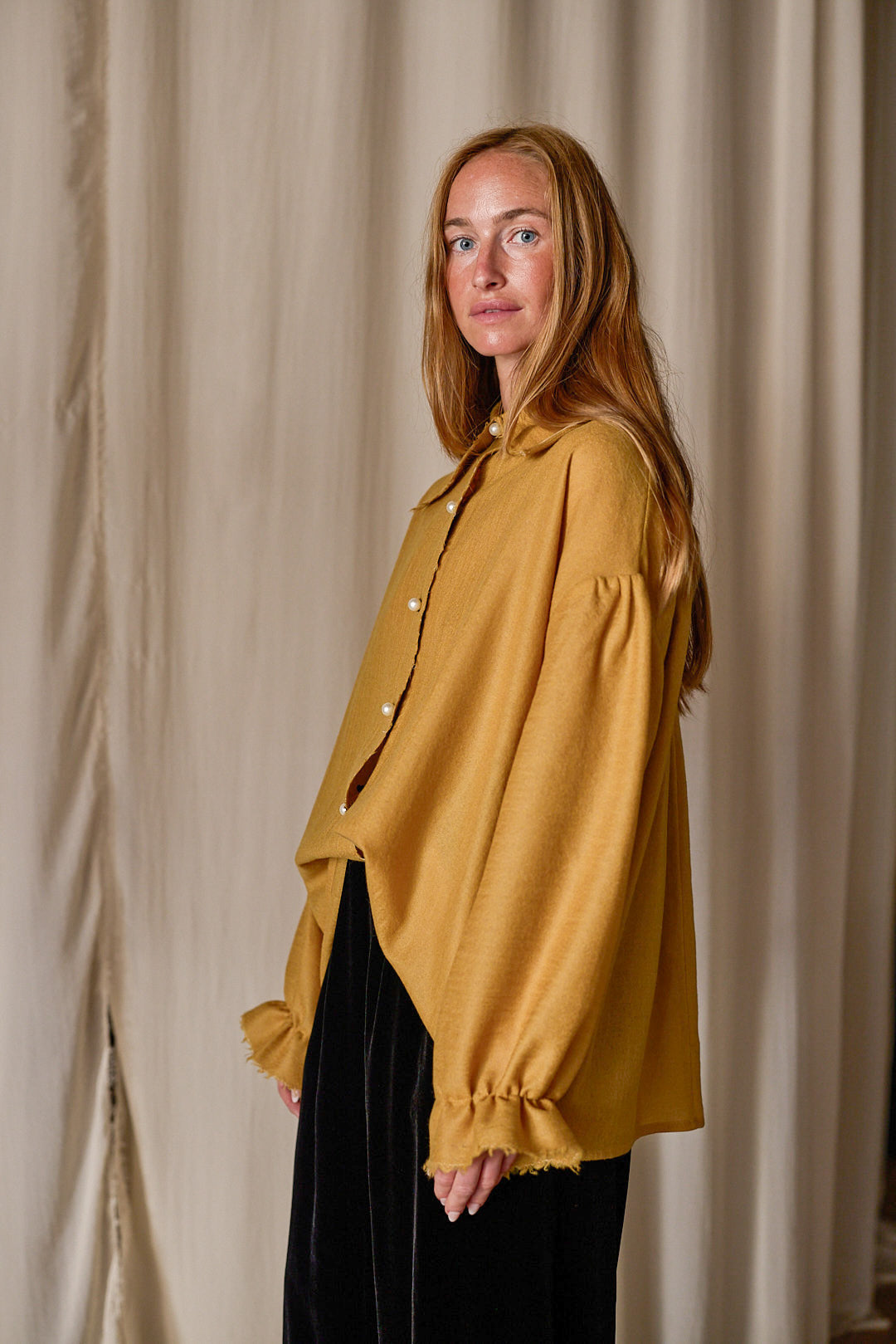 A woman with long, reddish hair stands against a beige curtain backdrop. She is wearing a mustard-yellow Poet Shirt | Japanese Wool Gauze Gold with billowy sleeves and black pants. She gazes calmly at the camera with a relaxed posture.